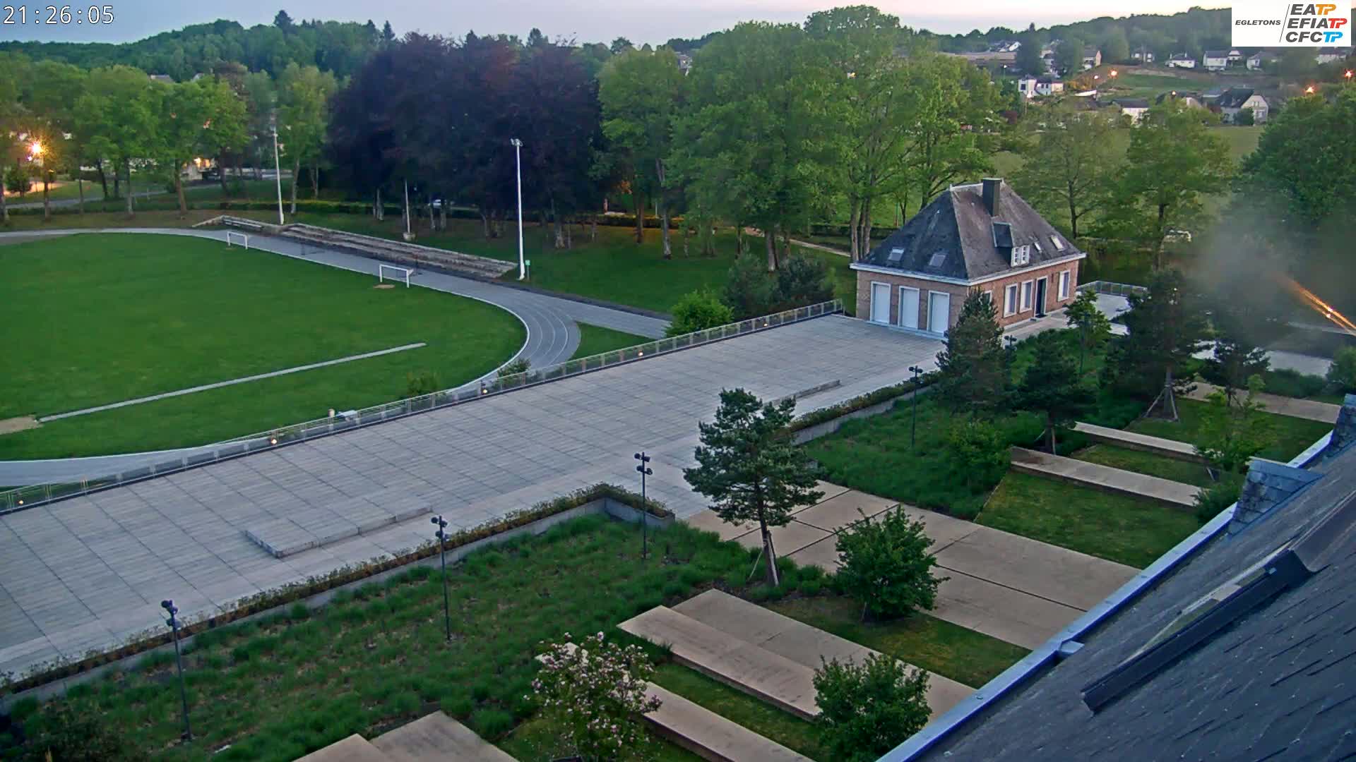 An aerial view shows a paved area with a running track and grass field, a small brick building, and landscaped gardens, all under a twilight sky.