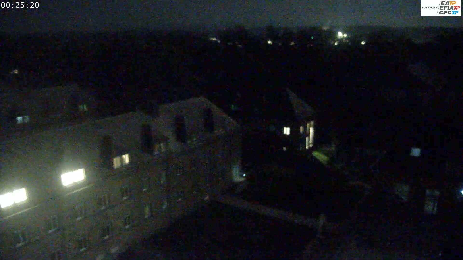 A nighttime, high-angle view shows several multi-story buildings with a few illuminated windows, surrounded by dark trees and other structures.