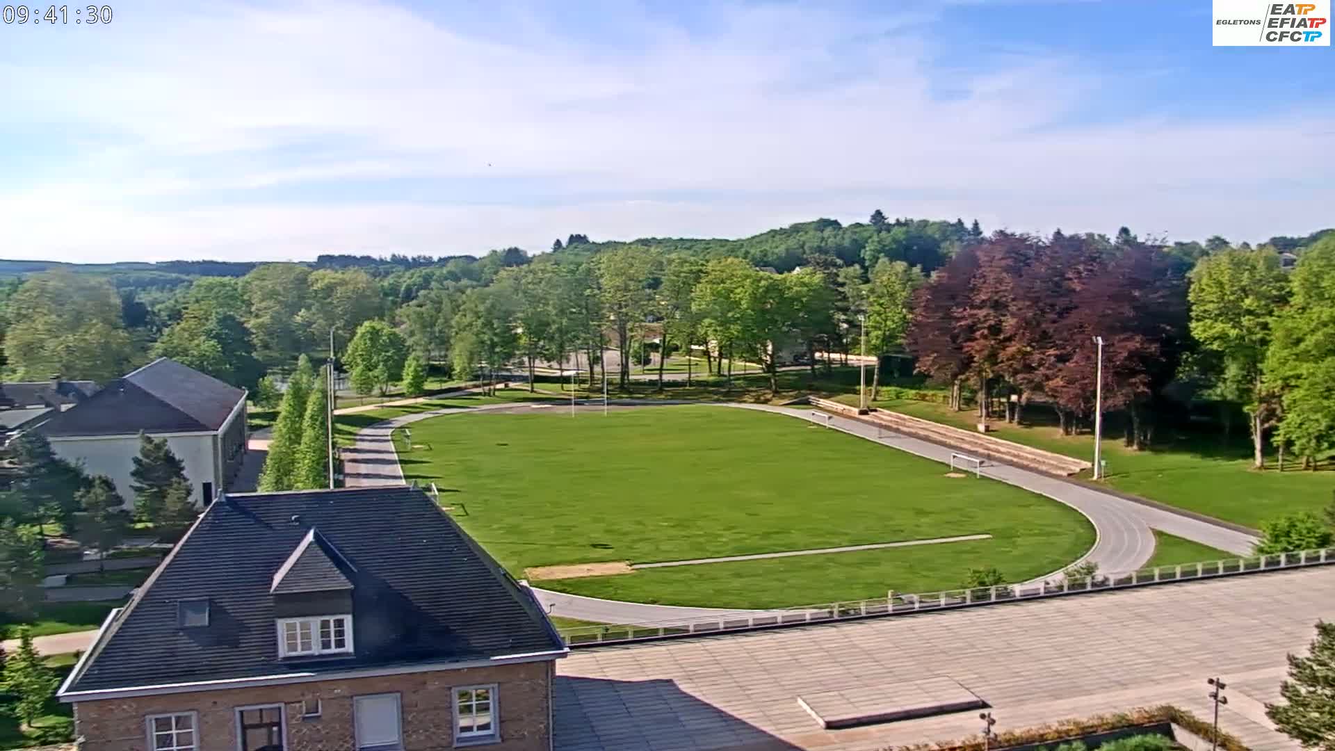 An aerial view shows a grassy athletic field with a running track, surrounded by trees and buildings under a mostly clear sky.