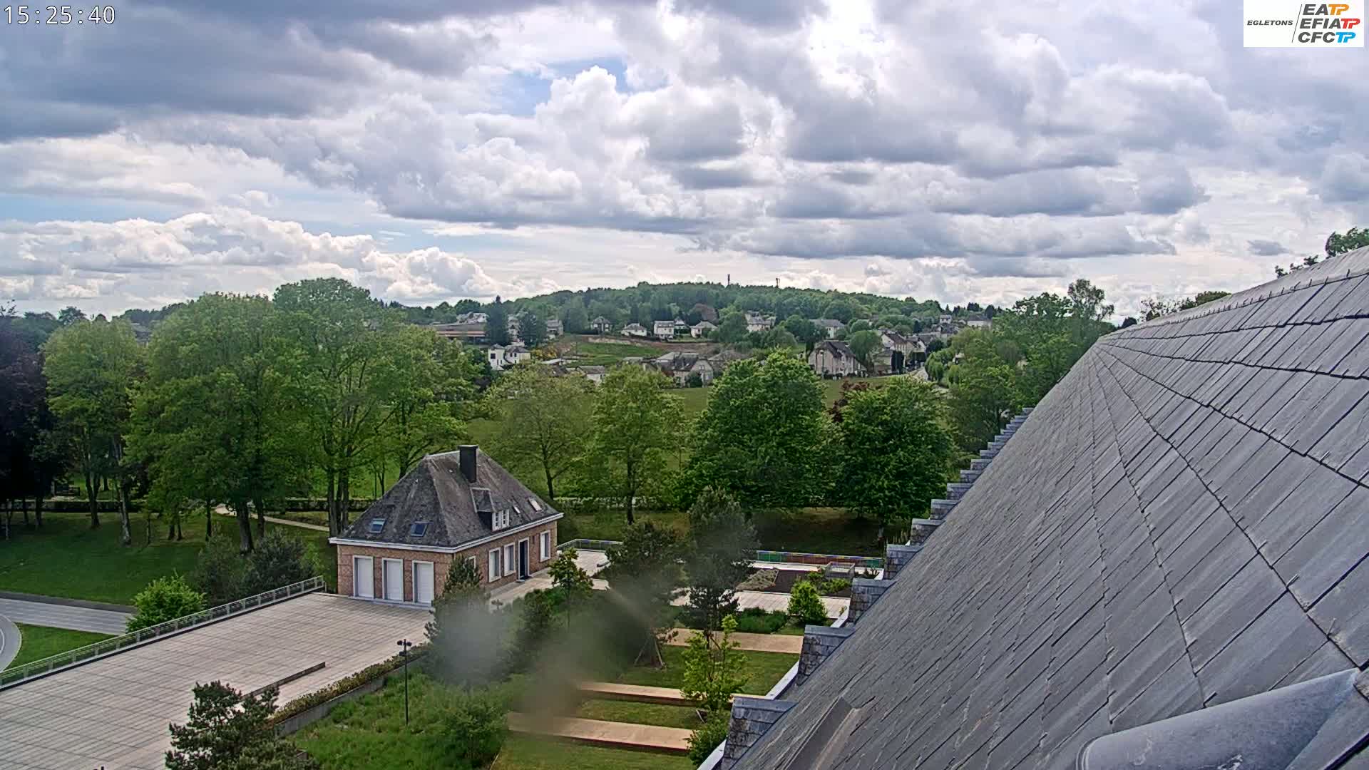 A partly cloudy sky overlooks a green landscape with a small brick building, a large sloped roof, and a distant residential area.