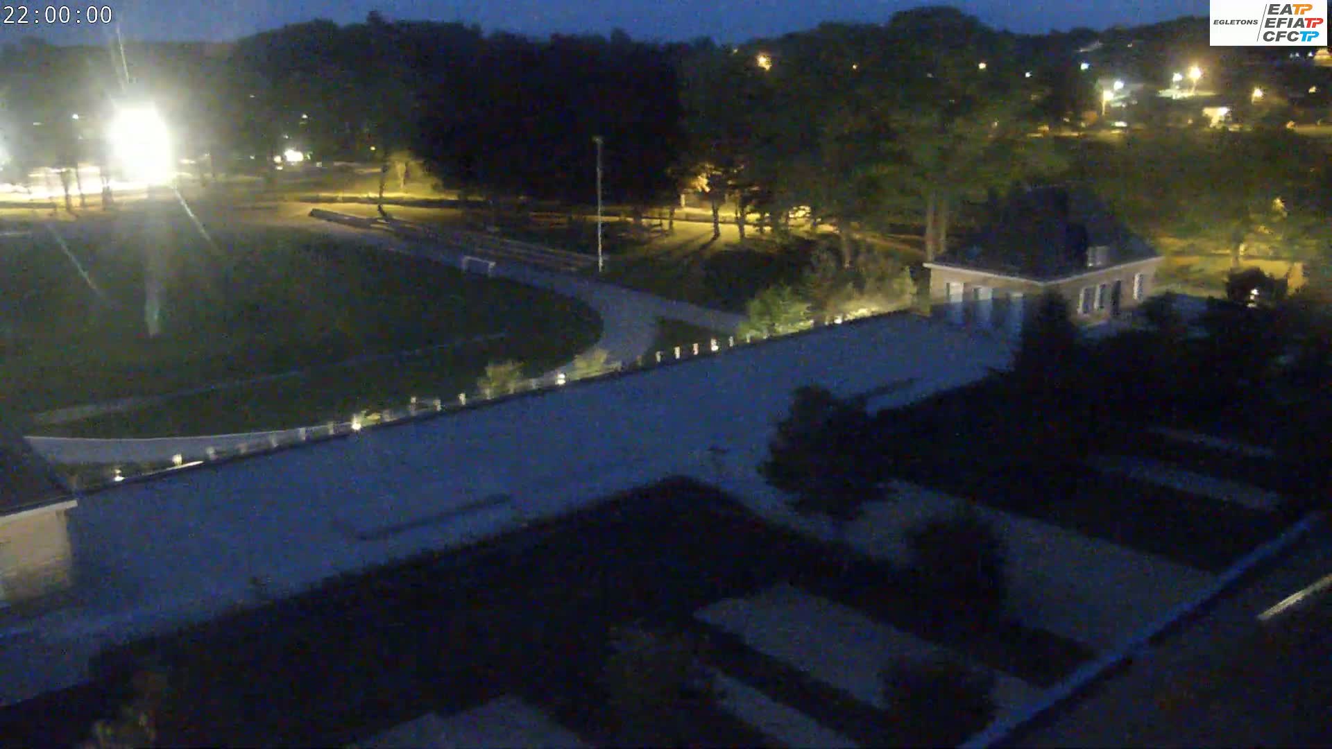 A nighttime aerial view shows a lit-up athletic field, surrounding trees, and a small building, all under a dark sky.