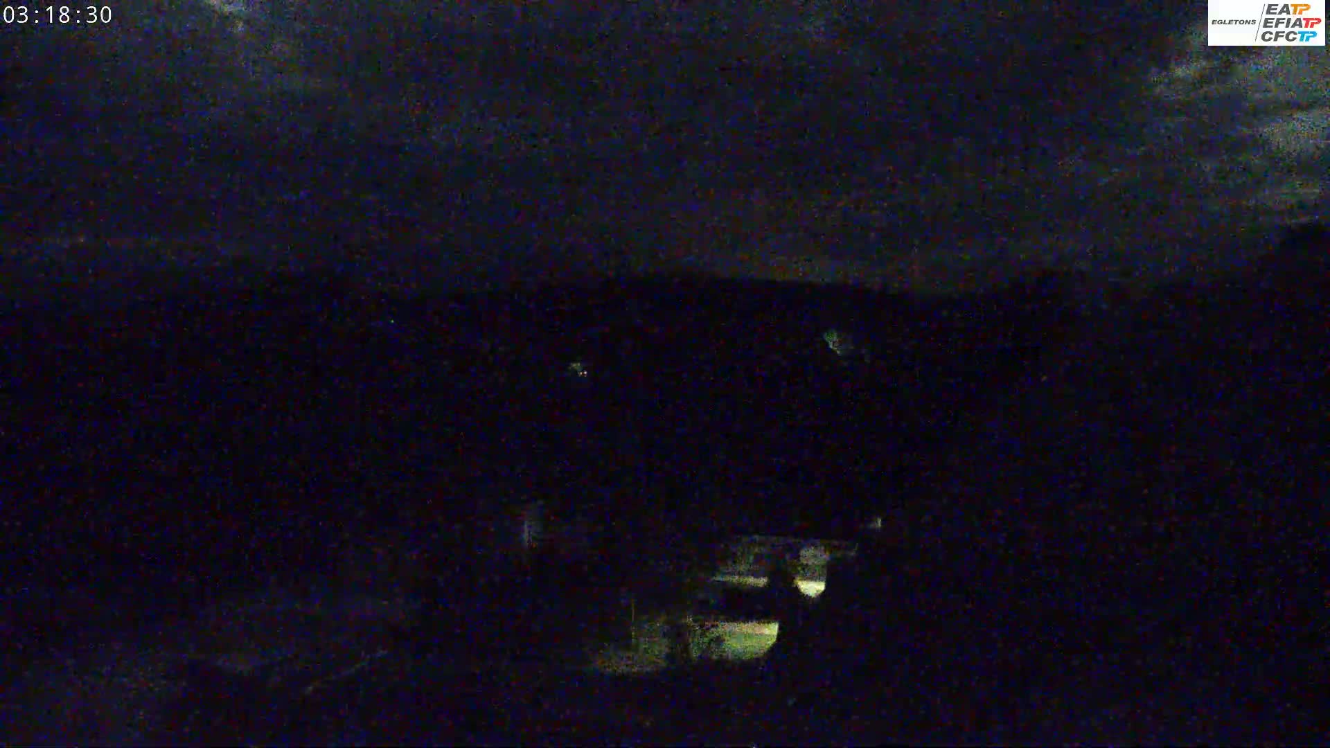 A nighttime view shows a dark landscape under a cloudy sky with a few faint lights visible in the distance.