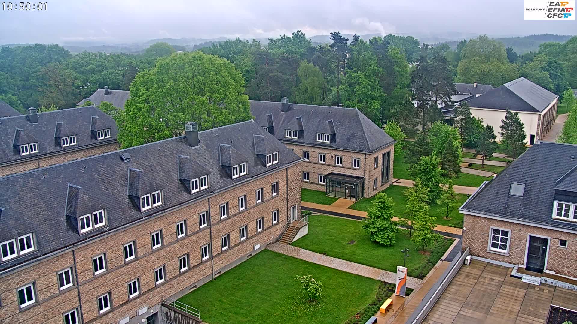 A high-angle view shows several low-rise brick buildings with slate roofs on a campus with lush green lawns and trees under an overcast sky.