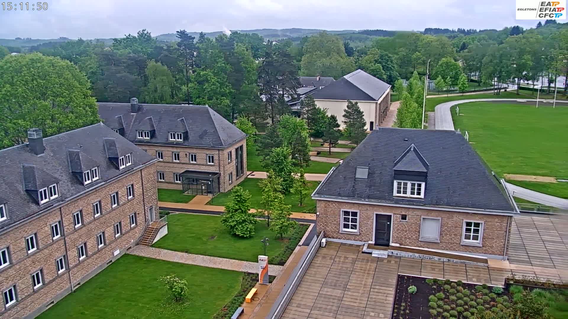 An aerial view shows several stone buildings on a campus with green lawns and trees under an overcast sky.