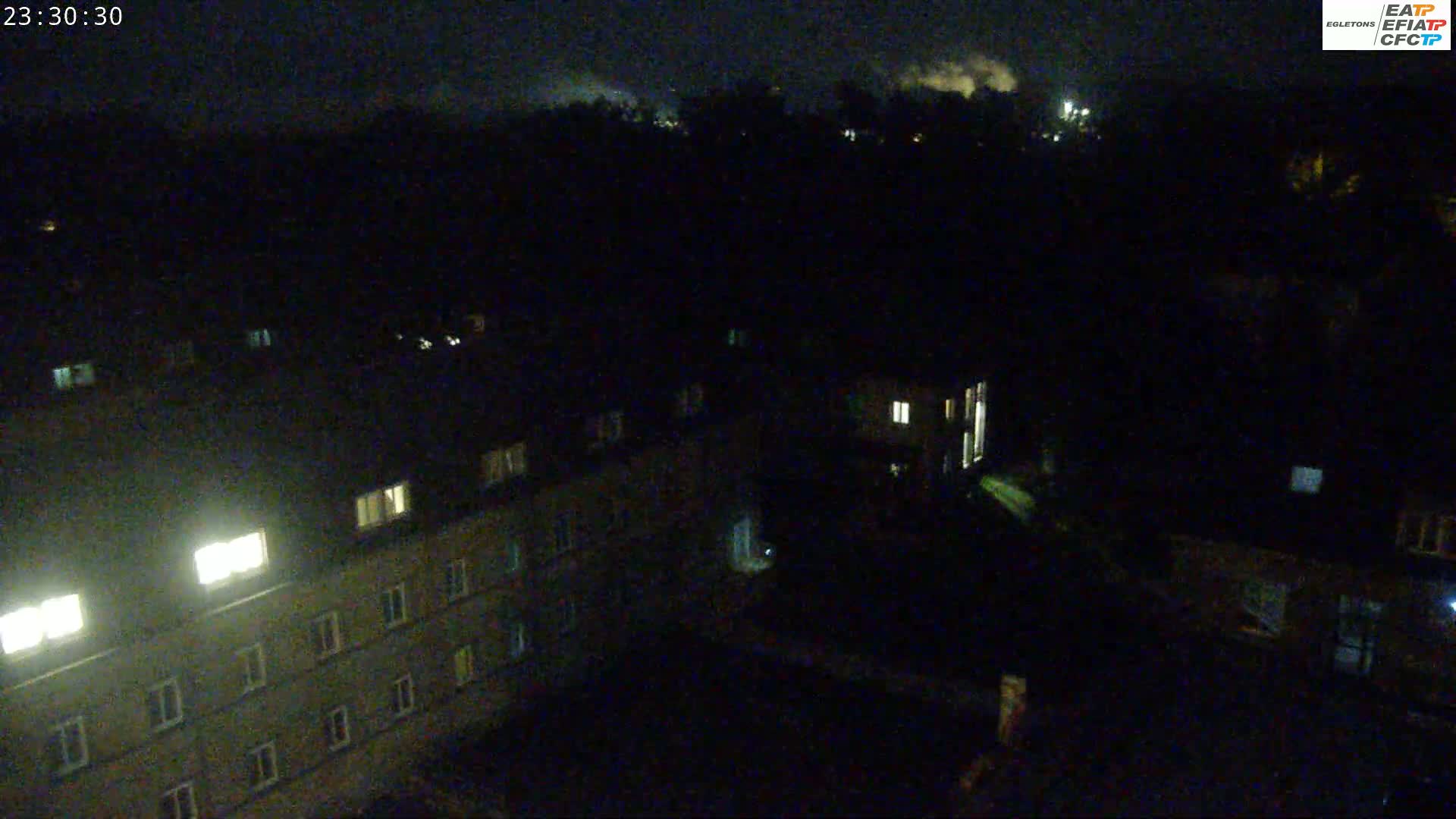 A nighttime, high-angle view of a dark residential area with some illuminated windows, and a slightly hazy sky.