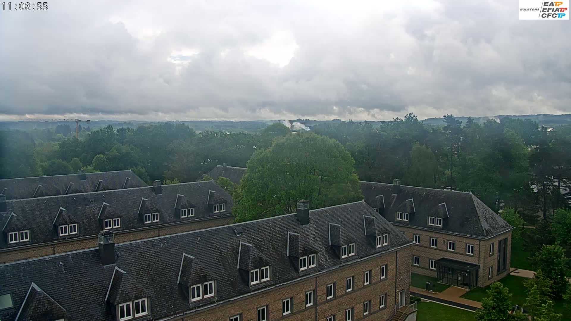 A cloudy day view of a large brick building with dark gray roofs, surrounded by lush green trees and a hazy distant landscape.