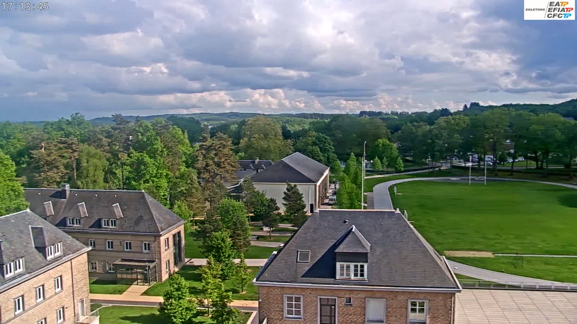 A high-angle view shows a partly cloudy day over a campus with several buildings, a large grassy field, and a running track, all surrounded by lush green trees.