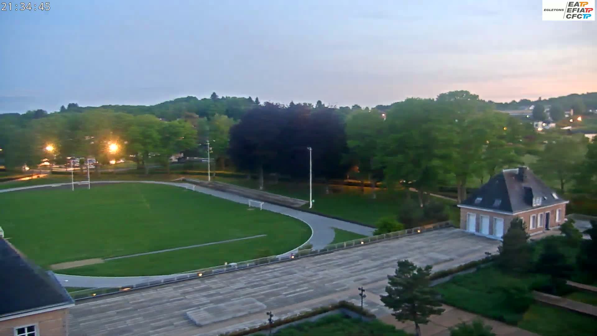 An aerial view shows a grassy athletic field with a running track surrounding it, adjacent to a paved area and a small brick building, all set within a landscape of trees under a twilight sky.