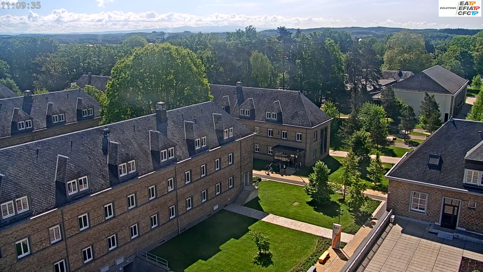 A high-angle view shows several low, long, stone buildings with dark gray roofs on a sunny day, surrounded by green lawns and trees, with distant hills visible in the background.