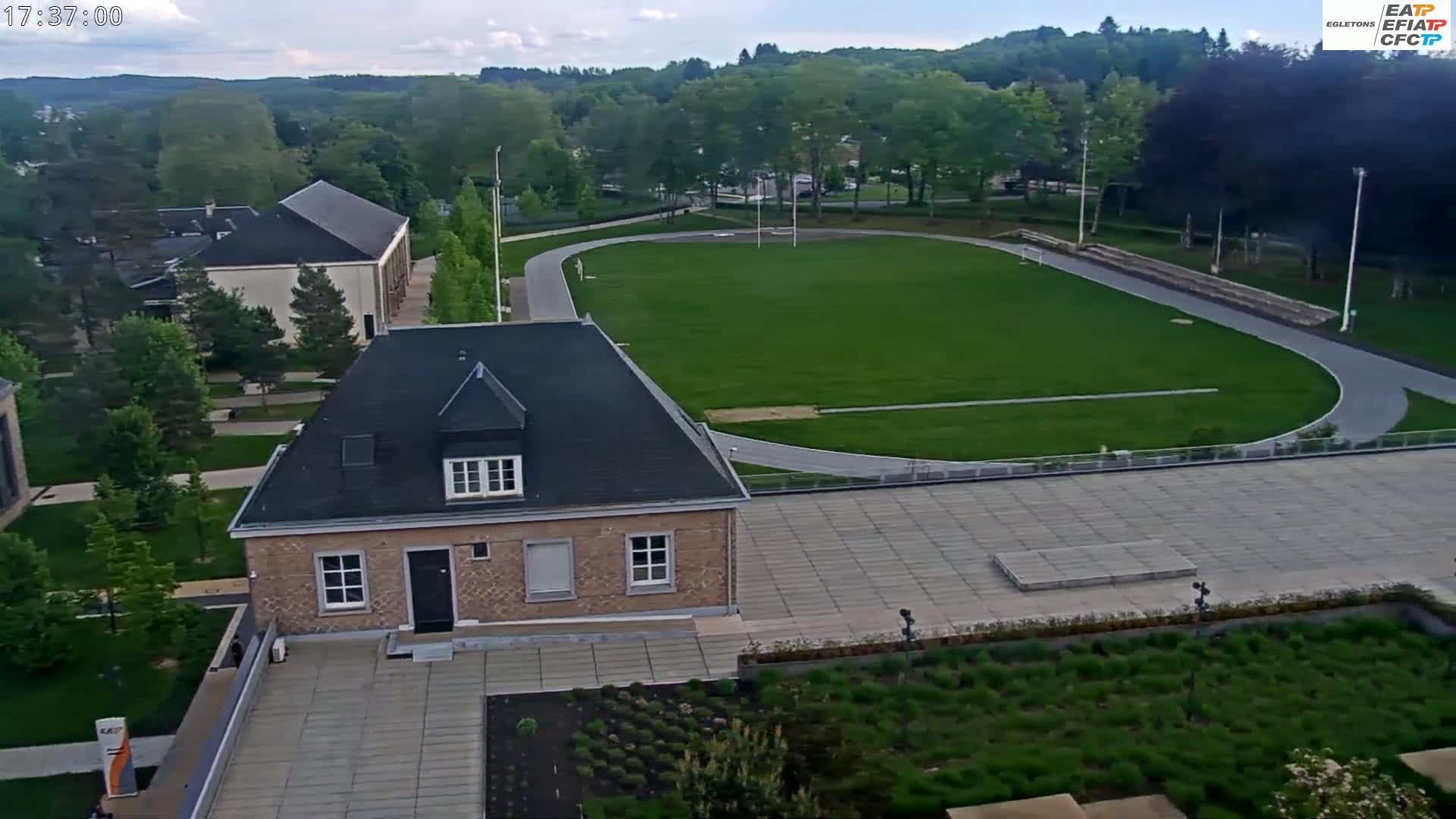An aerial view shows a small brick building next to a paved area and a grassy athletic field with a running track under a partly cloudy sky.