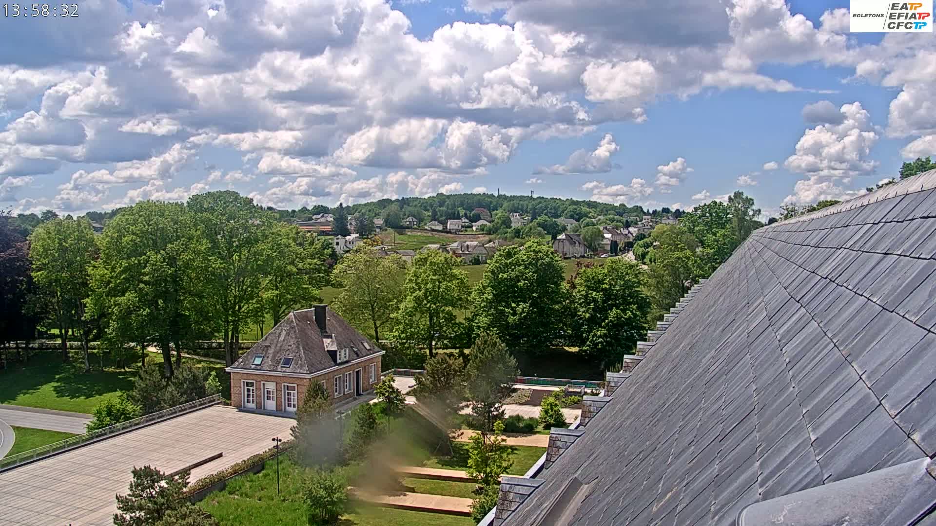 A partly sunny day reveals a view of a small town nestled among green hills and trees, as seen from a high vantage point, with a portion of a building's roof in the foreground.