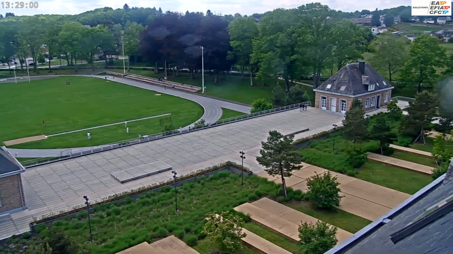 An aerial view shows a sports field, running track, paved walkways, a small building, and landscaped gardens on a partly sunny day.