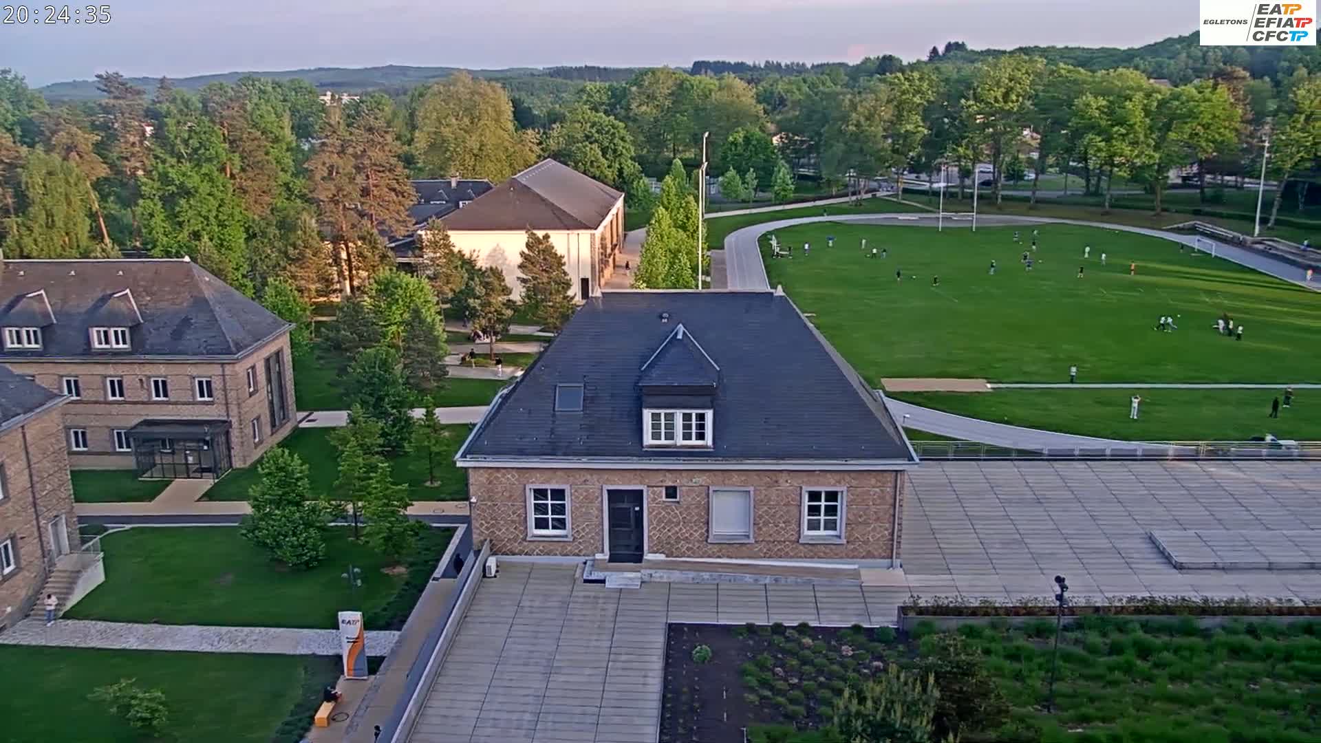 An aerial view of a campus at dusk shows several buildings, a large grassy field with people playing sports, and paved pathways.