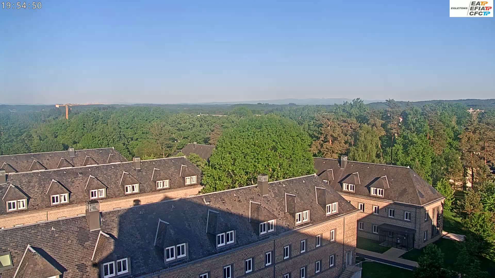 A high-angle view shows several brick buildings with dark gray roofs nestled amongst a lush green forest under a clear blue sky.
