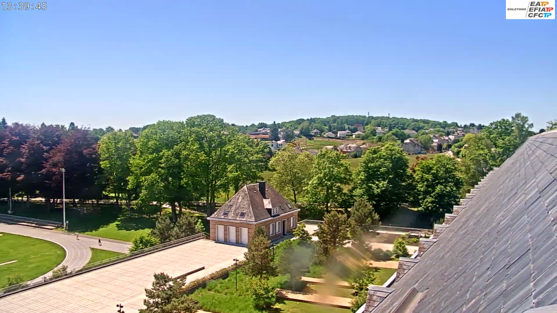 A sunny view from a rooftop shows a small building surrounded by landscaping, a running track, and a village nestled in a green hillside in the distance.