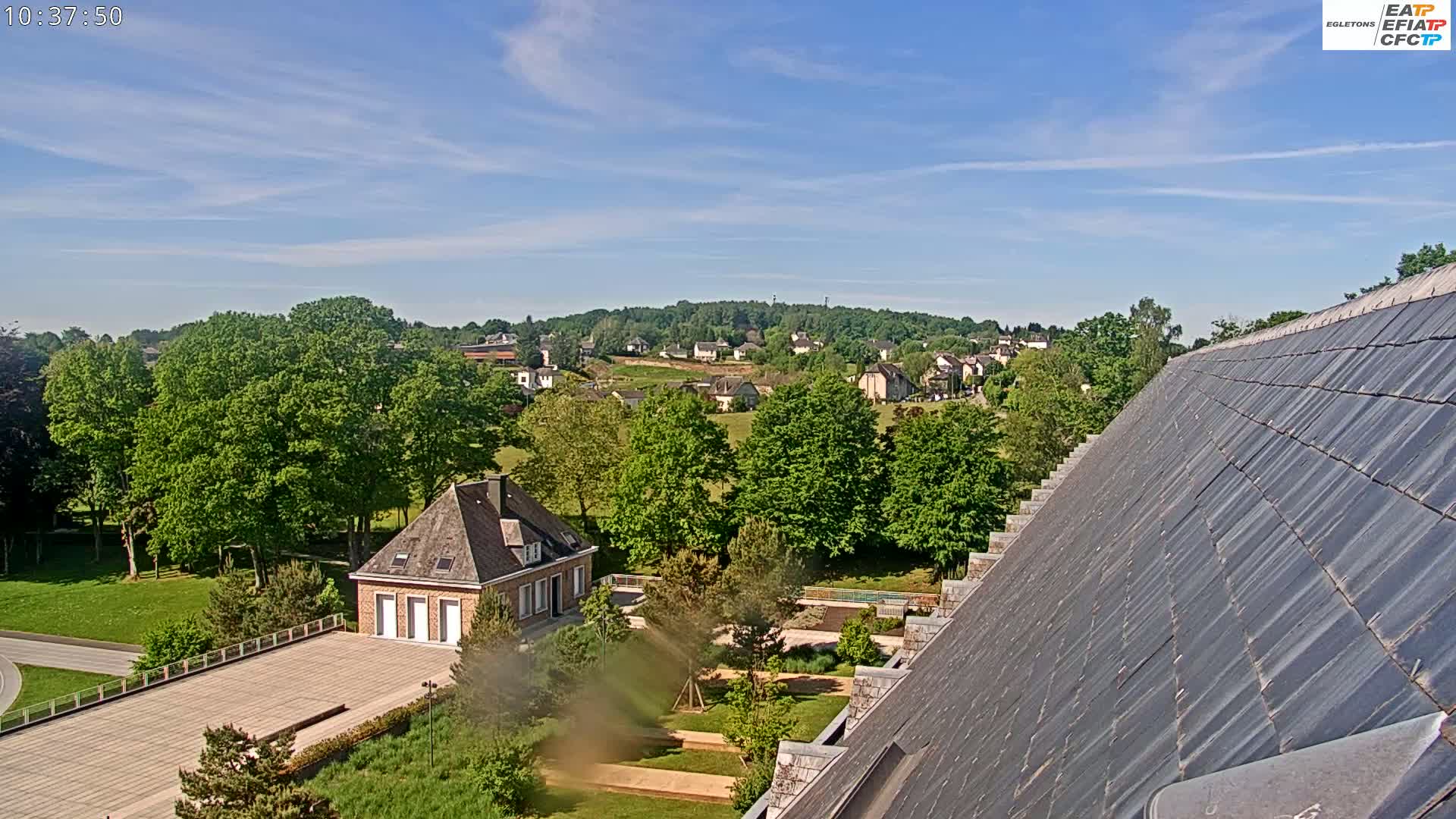 A partly sunny view from a high vantage point shows a slate roof in the foreground, a small brick building and manicured gardens below, and a village nestled in a valley beyond.