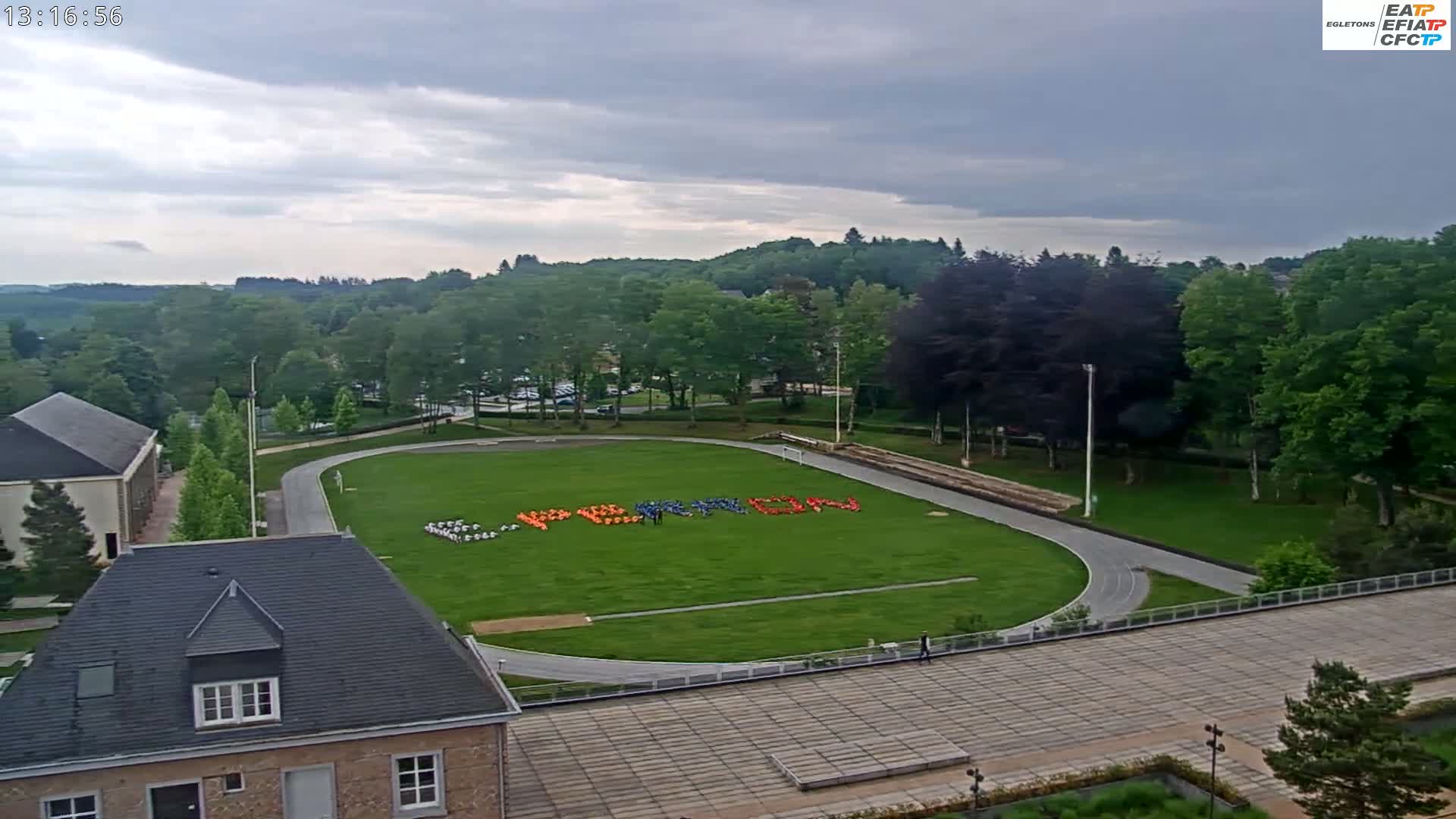 An aerial view shows a grassy field with a running track surrounding it, people forming a word on the field, and a partly cloudy sky.