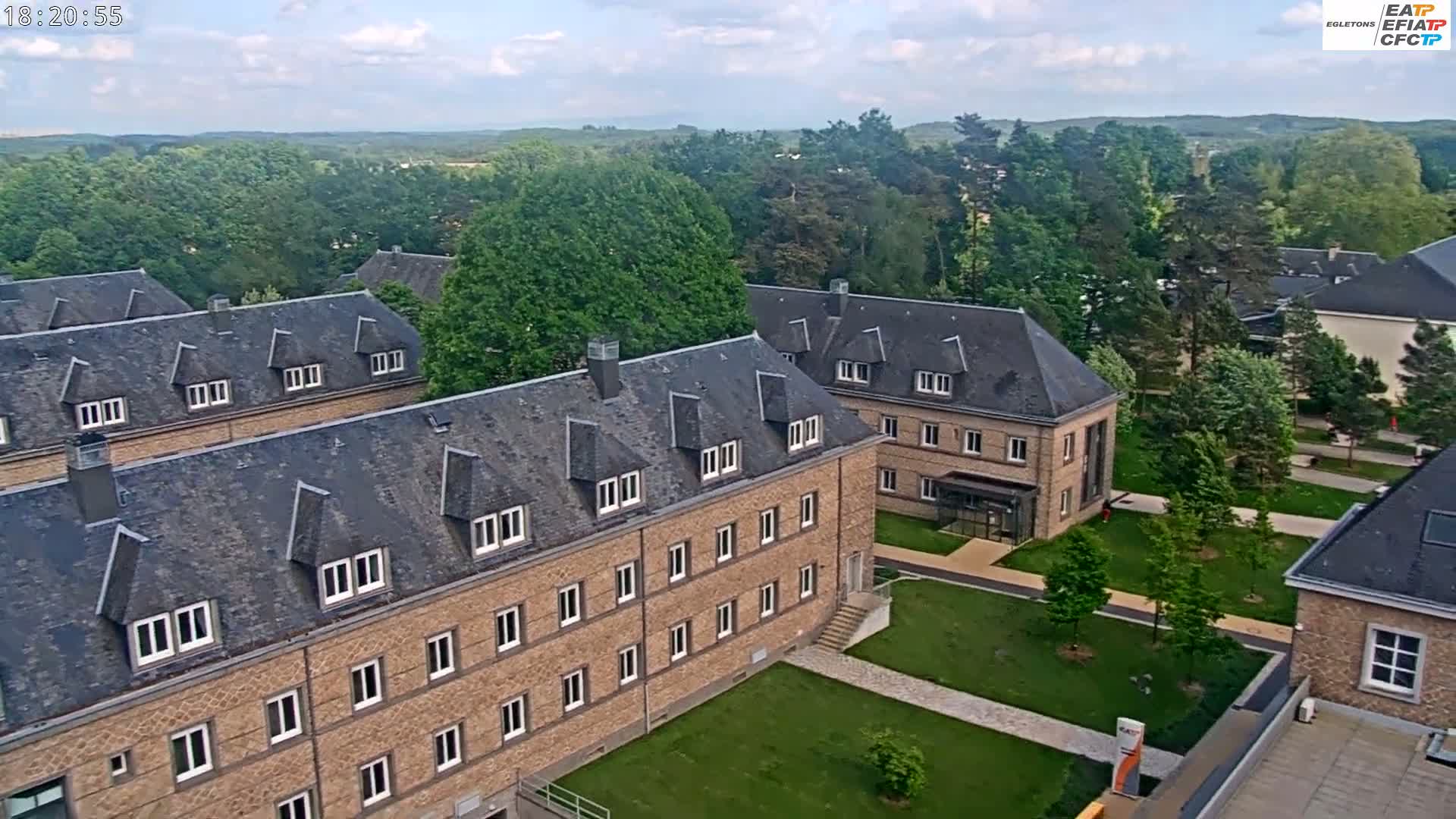 An aerial view shows several multi-story brick buildings with slate roofs, surrounded by green lawns and trees under a partly cloudy sky.