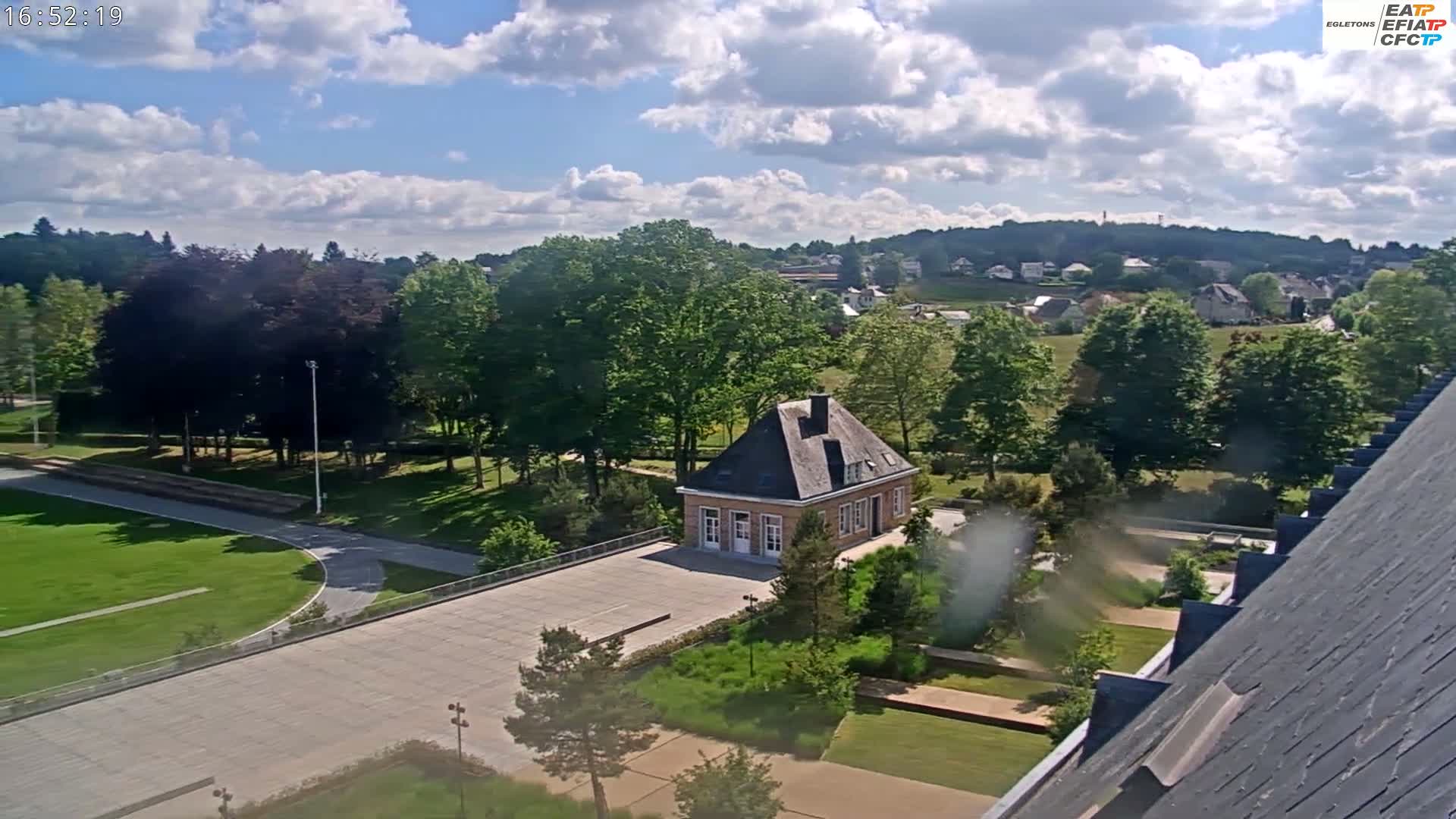 A partly cloudy day reveals a landscape with a small stone building situated in a courtyard amidst lush green trees, manicured lawns, and paved pathways, overlooking a residential area on a hillside.