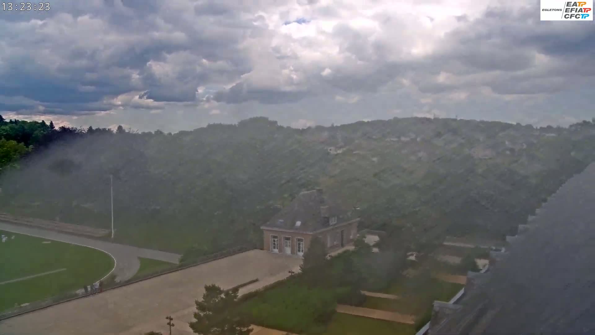 A hazy, overcast day reveals a landscape of green hills, a small stone building in a courtyard, and a grassy field with a track, all viewed from an elevated perspective.