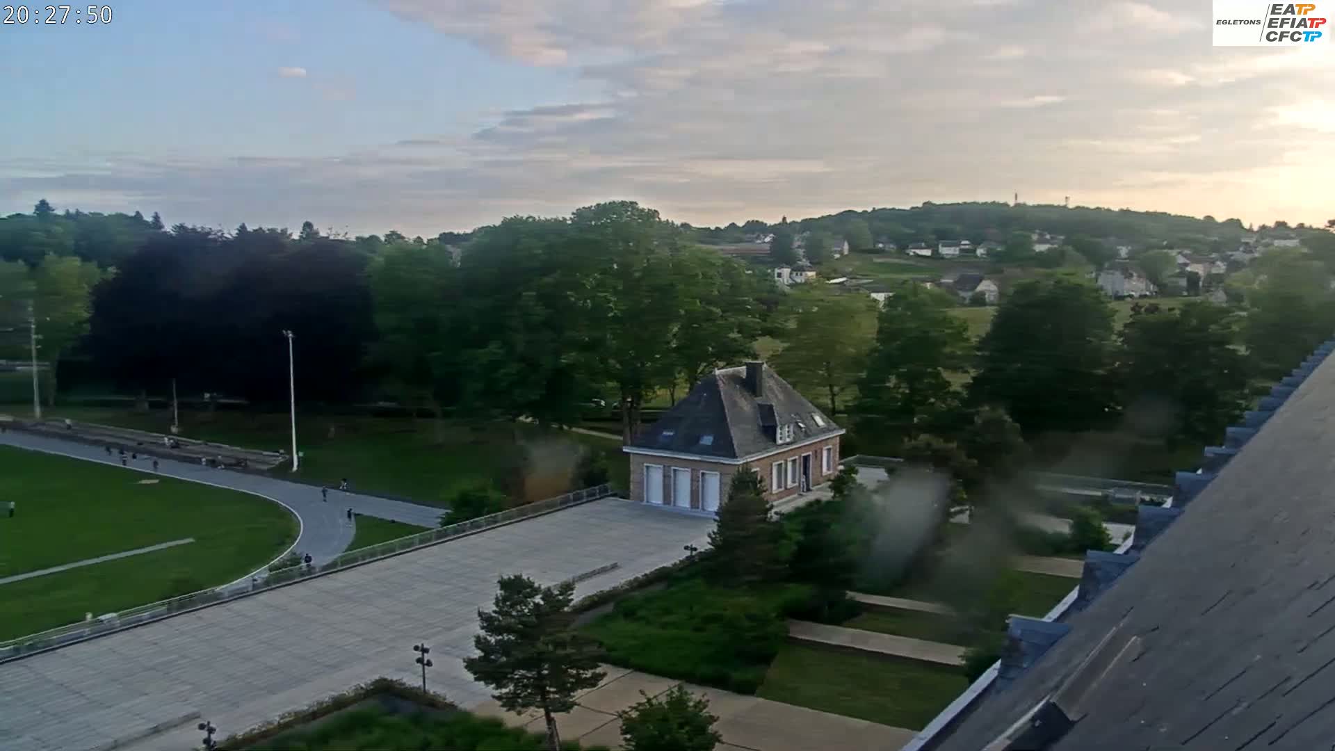 An elevated view shows a small, light-brown building on a paved courtyard, surrounded by green lawns, trees, and a track with people on it, all under a partly cloudy evening sky.