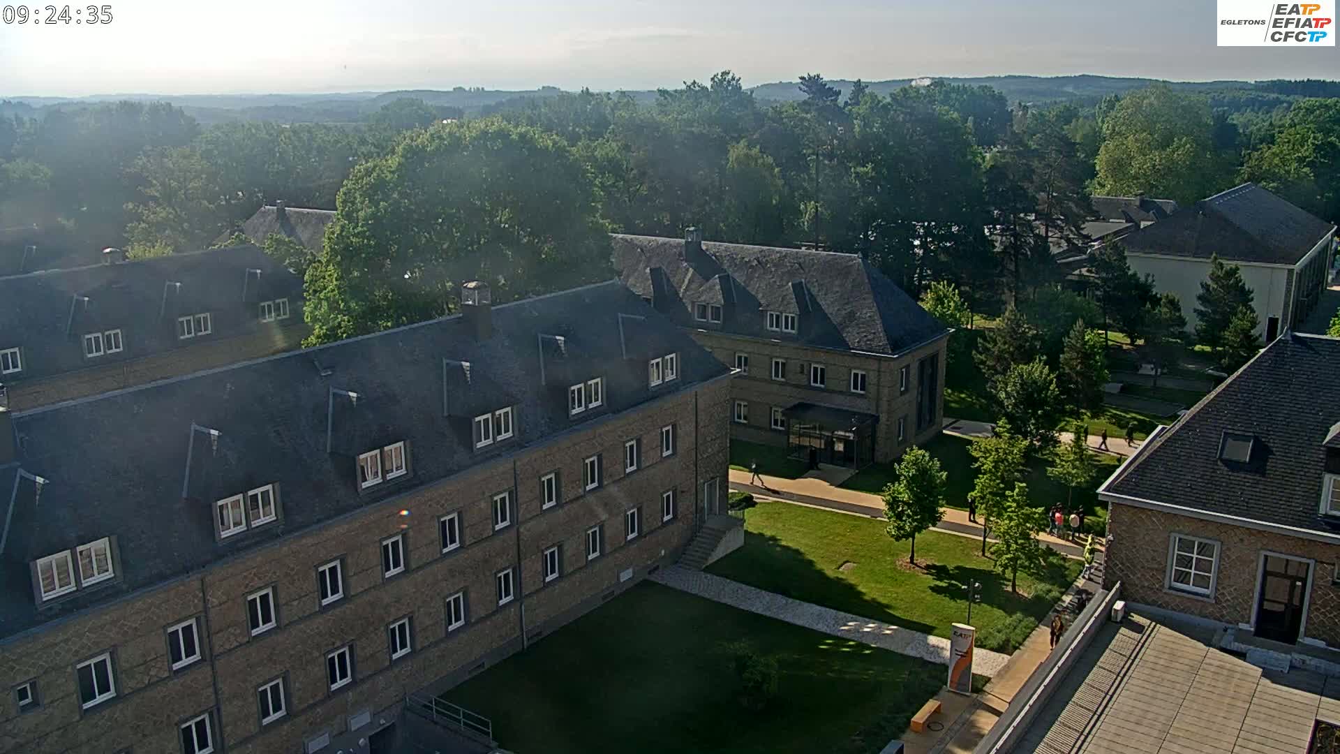 A hazy morning view of a campus with several brick buildings and green lawns, surrounded by trees and a distant line of hills.