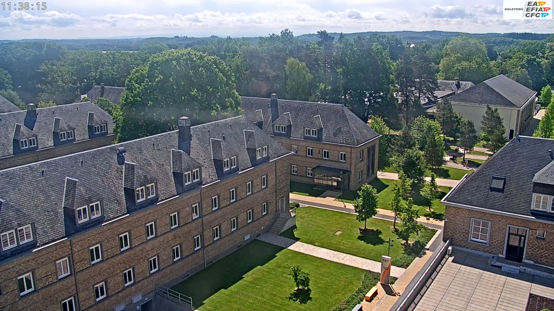 A high-angle view of several stone buildings on a sunny day, surrounded by green lawns and trees, with a forest visible in the background.