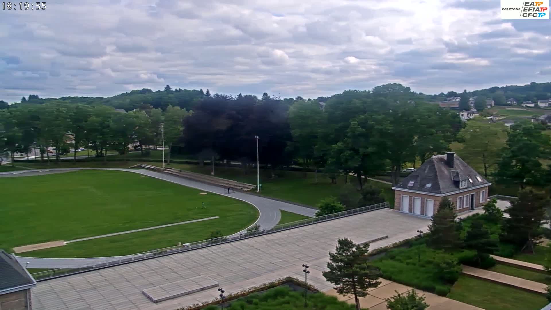 An aerial view shows a green grassy field with a running track, a small stone building, and surrounding trees under a partly cloudy sky.