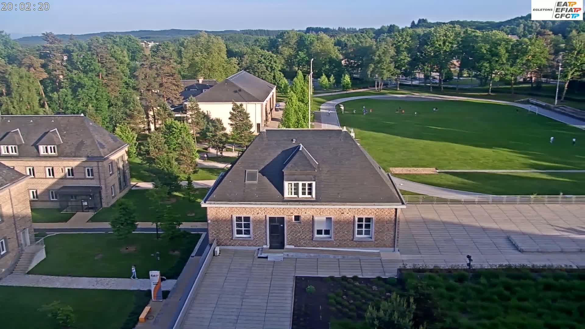 An aerial view shows several buildings on a campus with a large grassy field and people visible in the distance under a mostly clear sky.