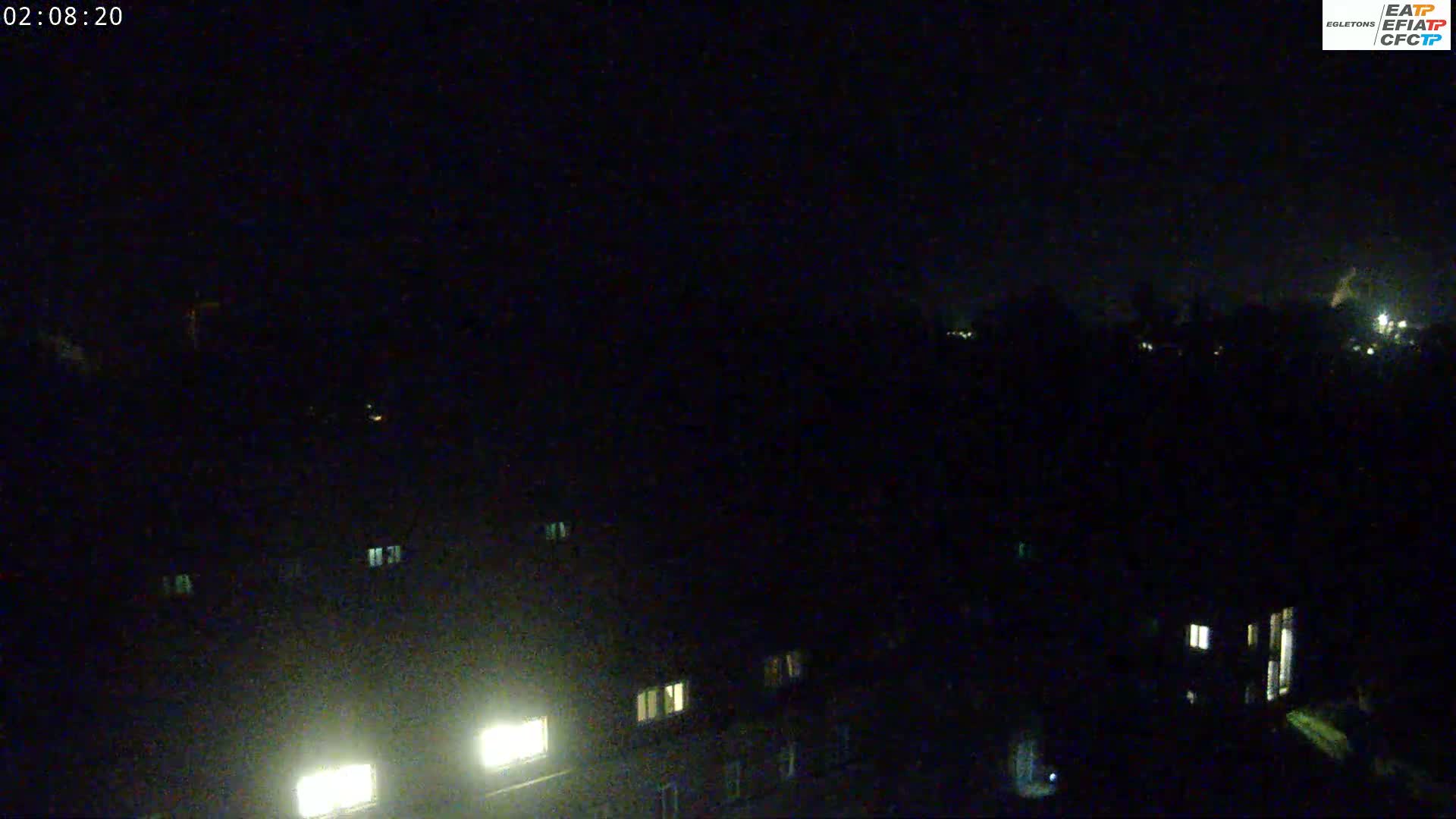 A nighttime, low-light view of buildings with illuminated windows, showing a dark, possibly overcast sky.