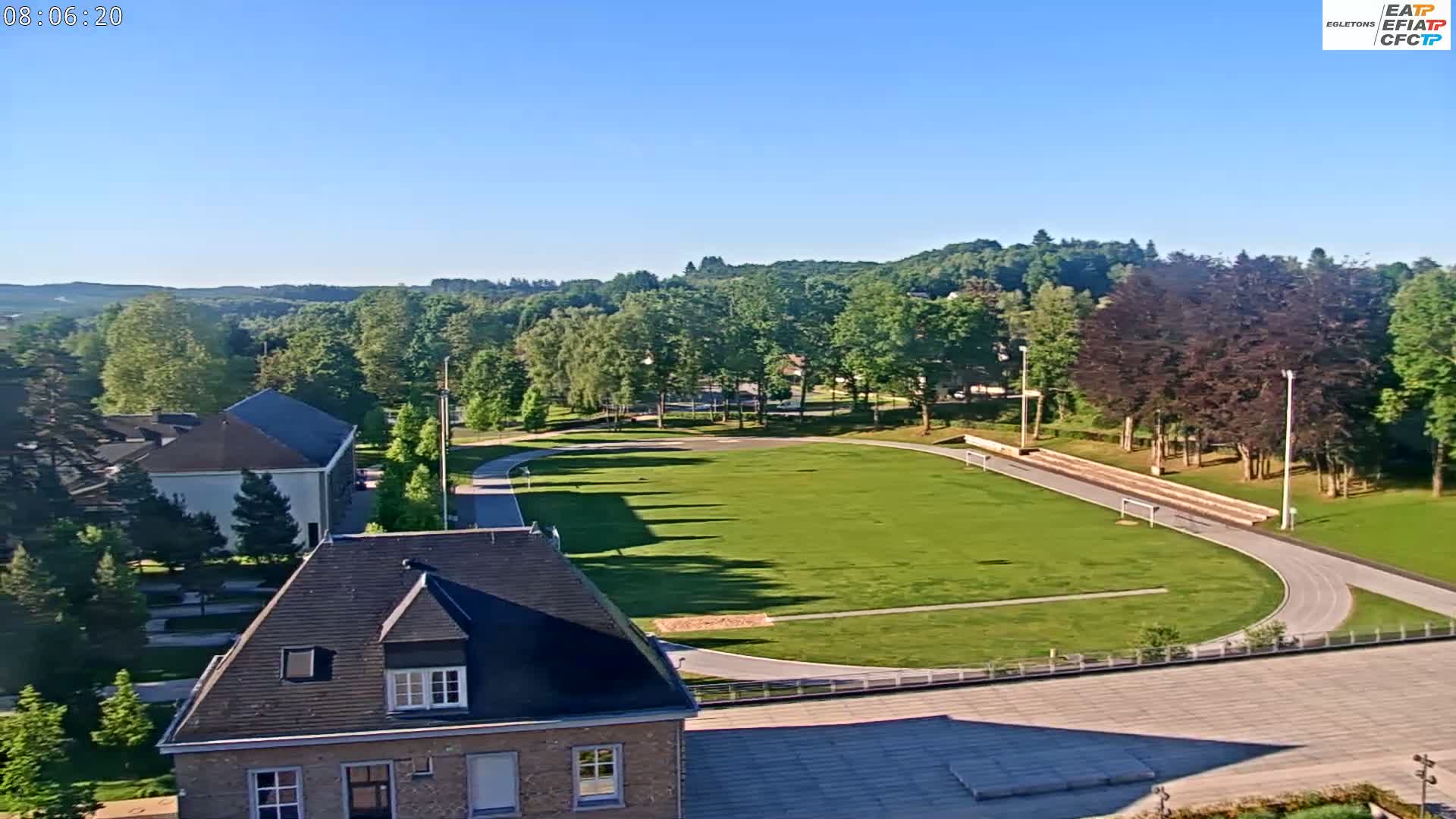 A high-angle view on a clear, sunny day shows a grassy field with a running track, bordered by trees and buildings.