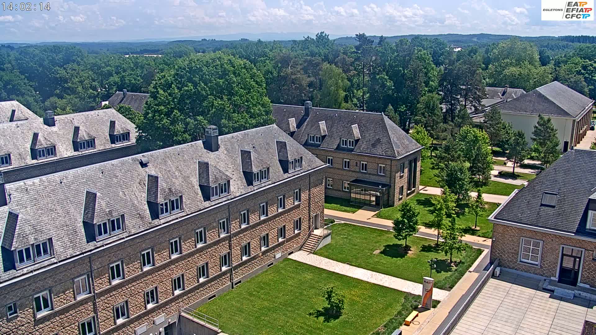 An aerial view of a campus on a sunny day shows several stone buildings surrounded by green lawns and trees, with a distant view of rolling hills.