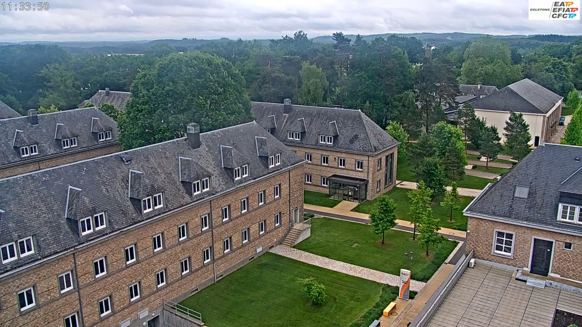 An aerial view shows several low, brick buildings with slate roofs, surrounded by lawns and trees under an overcast sky.