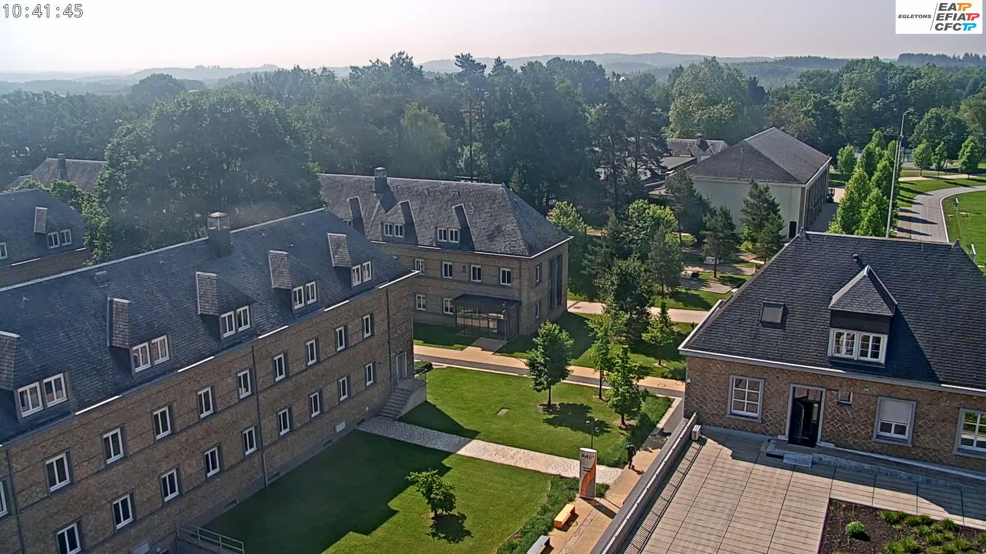 A high-angle, long shot on a hazy day shows several low, stone buildings with dark gray roofs on a campus-like setting with manicured lawns and trees.