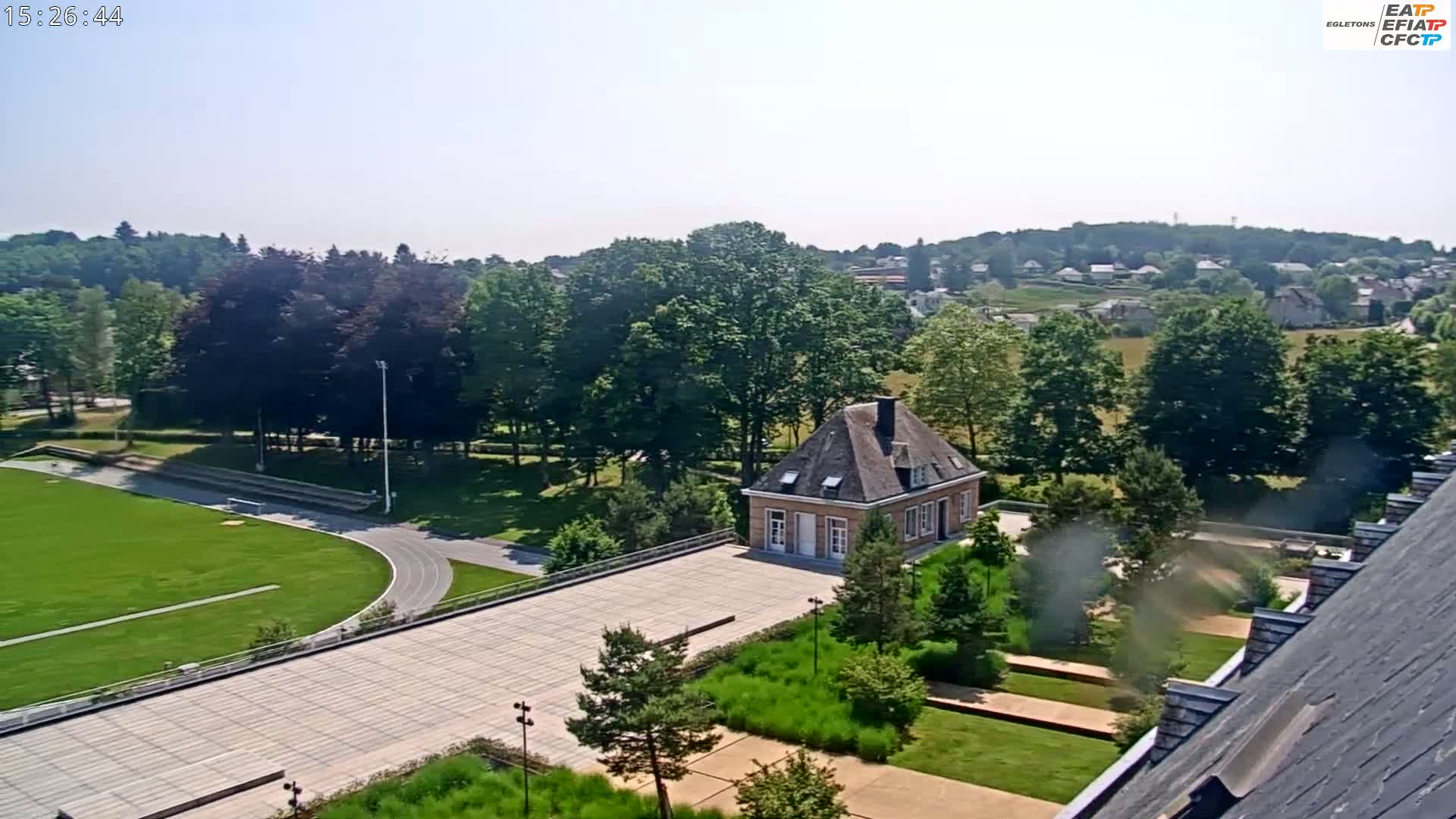 An aerial view shows a small, gabled building in a landscaped area with a running track and green fields, all under a clear, sunny sky.