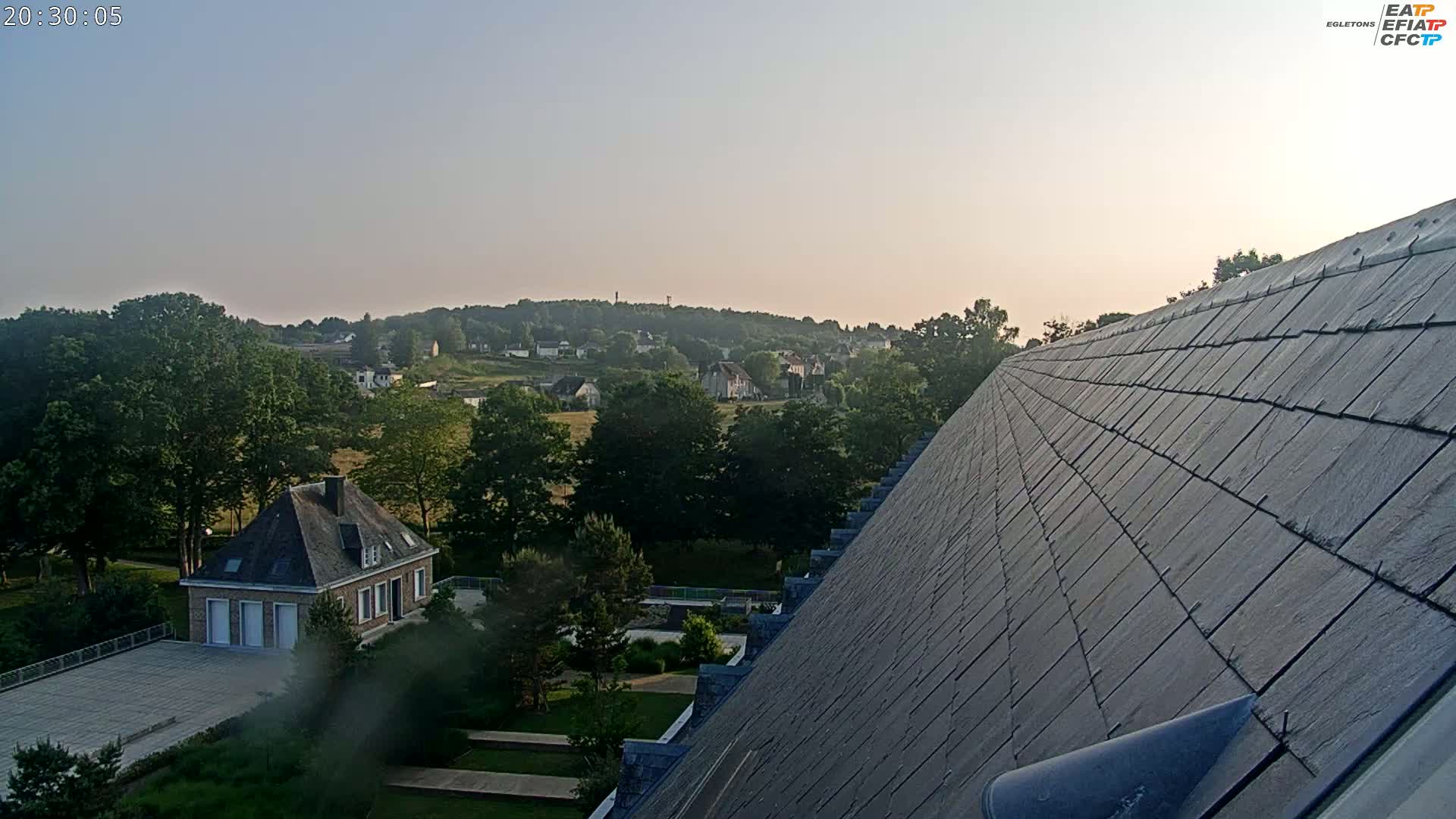 A view from a sloped roof shows a clear evening sky over a landscape of green trees, a flat field, and several light-colored buildings in a village.
