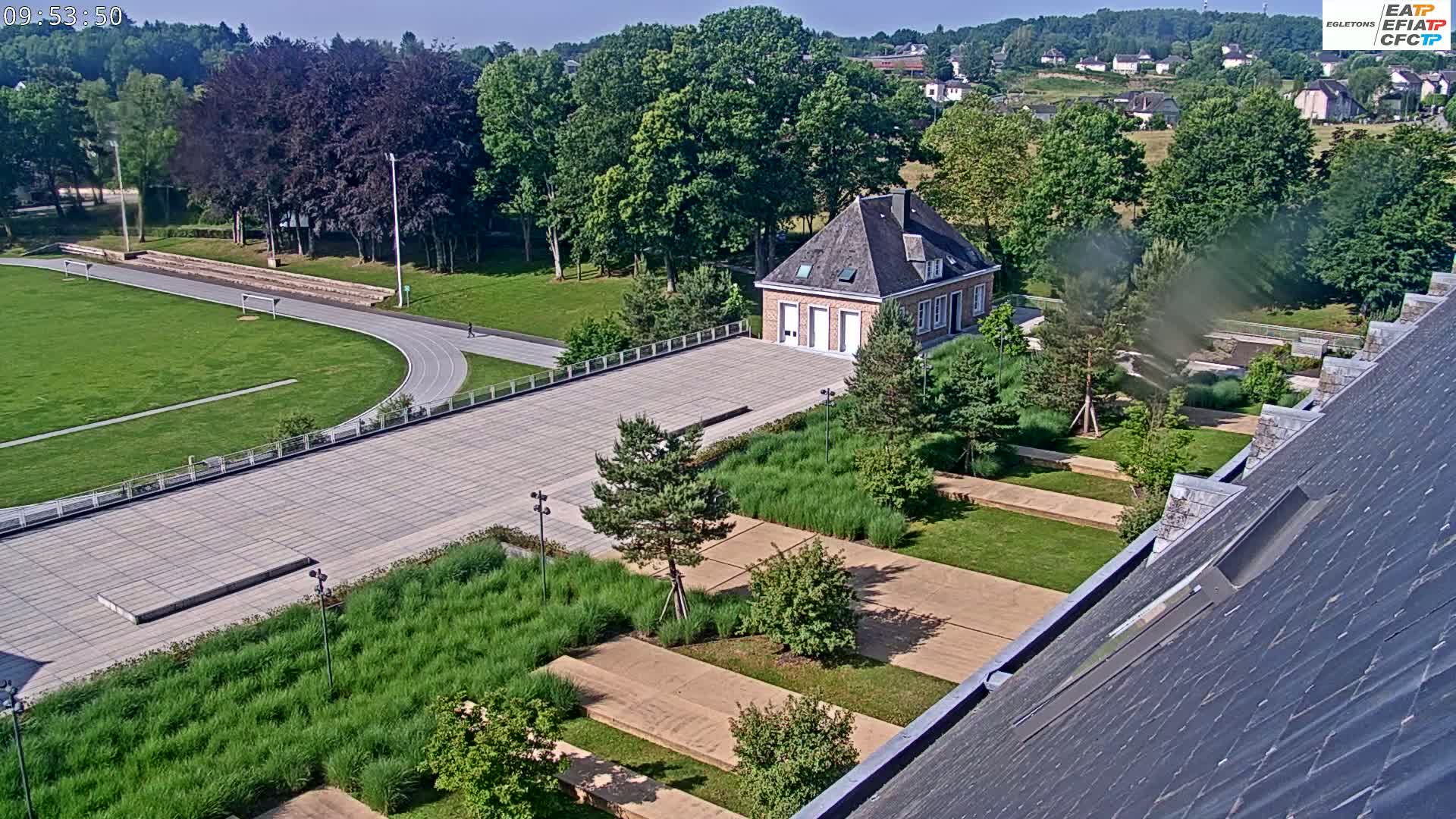 An aerial view shows a running track, a brick building, landscaped gardens with pathways, and a section of a dark-grey roof under a sunny sky.