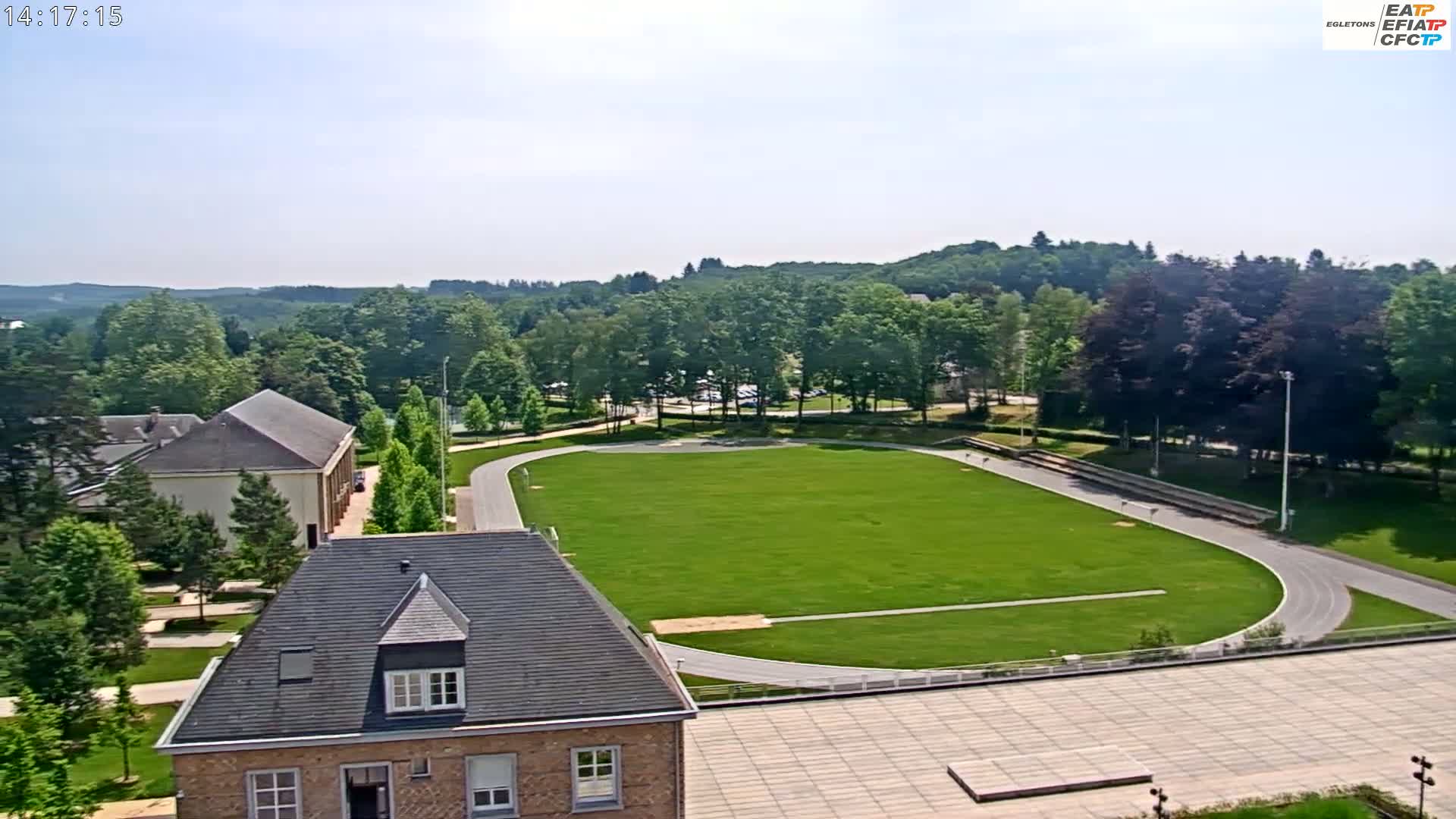 An aerial view shows a grassy athletic field with a running track, surrounded by trees and buildings under a mostly clear sky.