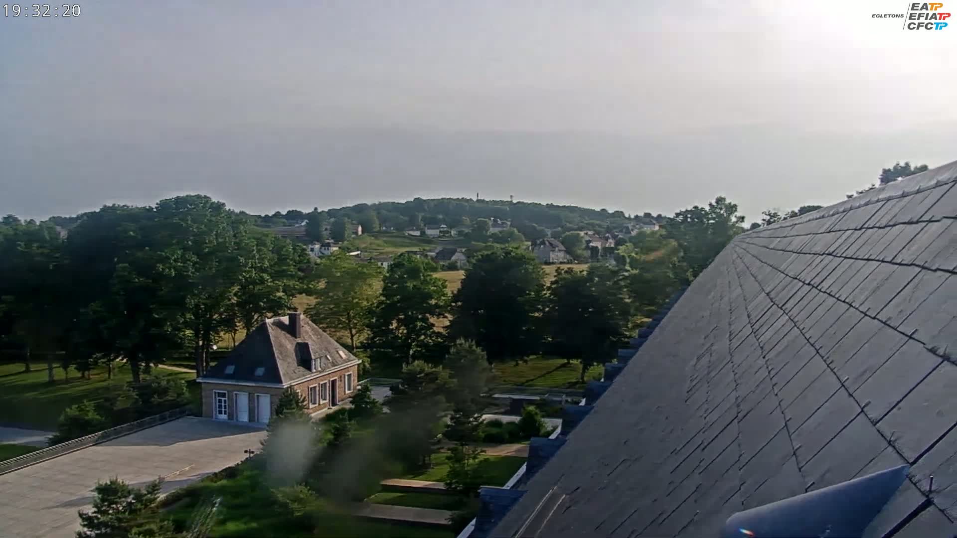 A hazy view from a sloped rooftop shows a small brick building and a sprawling green landscape with trees and distant houses under a mostly clear sky.