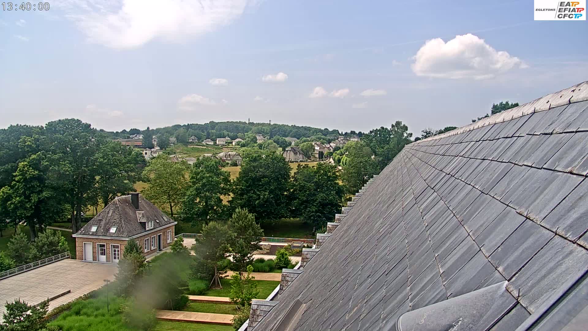 A partly cloudy sky overlooks a village nestled amongst green trees and fields, viewed from a steeply pitched slate roof.