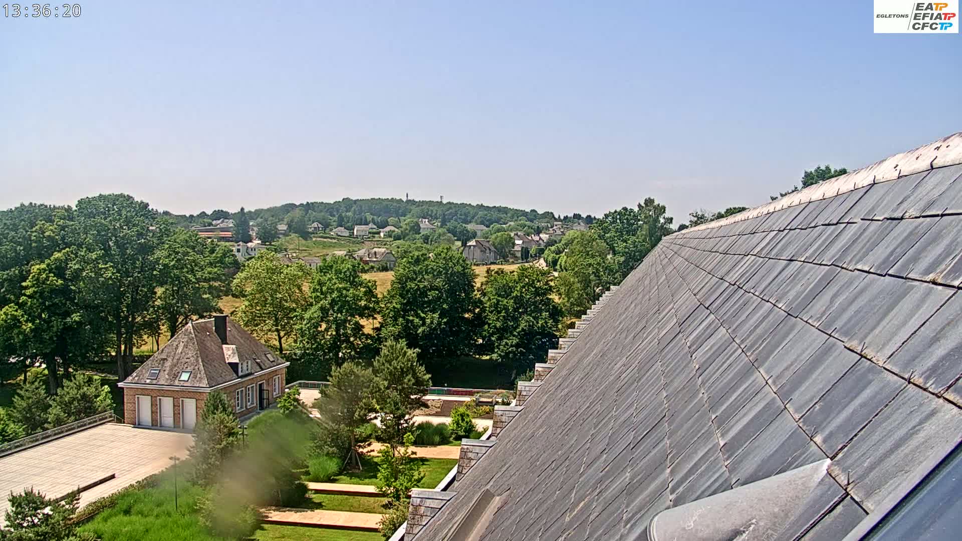 A sloped slate roof overlooks a landscape of green trees, a small village in the distance, and a manicured garden with a building below, all under a clear, sunny sky.