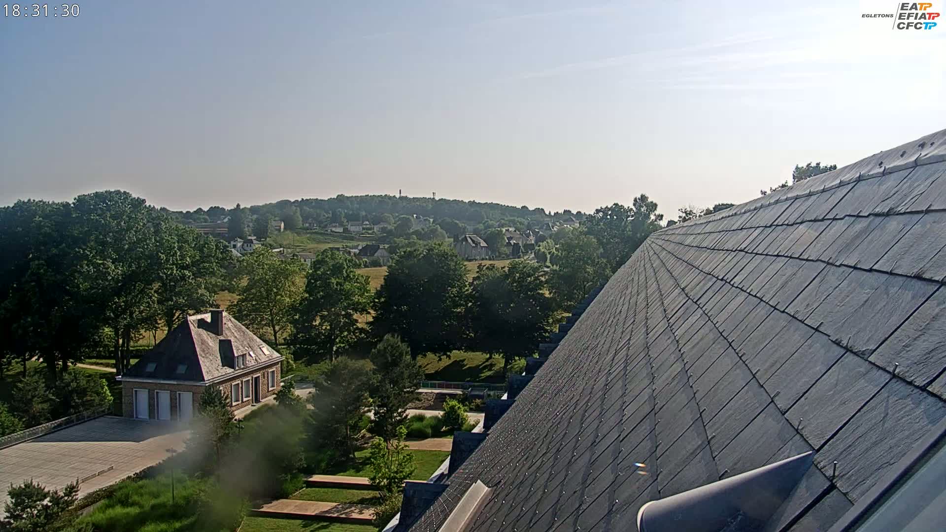 A sloped slate roof overlooks a tranquil landscape of green trees, a small village in the distance, and a brick house nestled within the greenery under a clear, sunny sky.