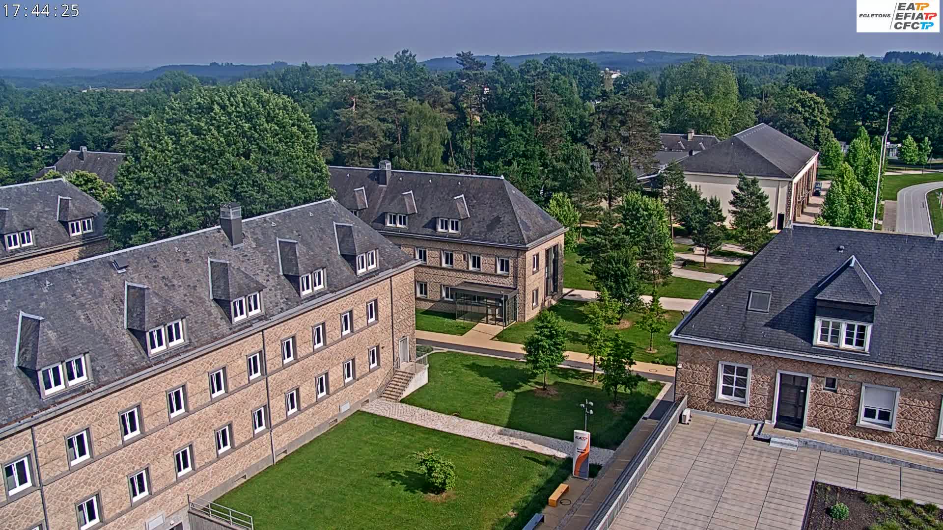 An aerial view of a campus of stone buildings on a sunny day, surrounded by lush green trees and lawns.