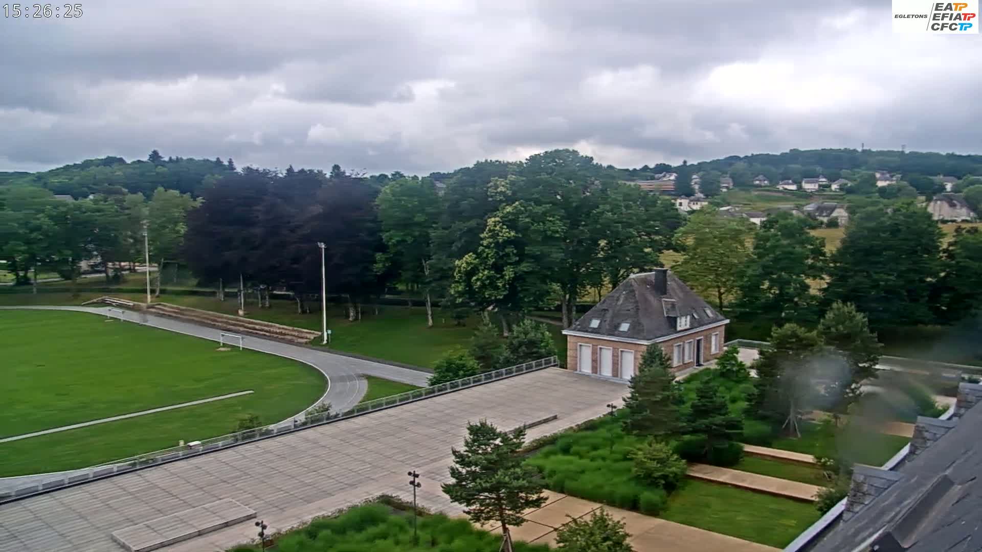 An aerial view shows a running track and grassy field surrounded by trees and a small brick building on a mostly cloudy day.