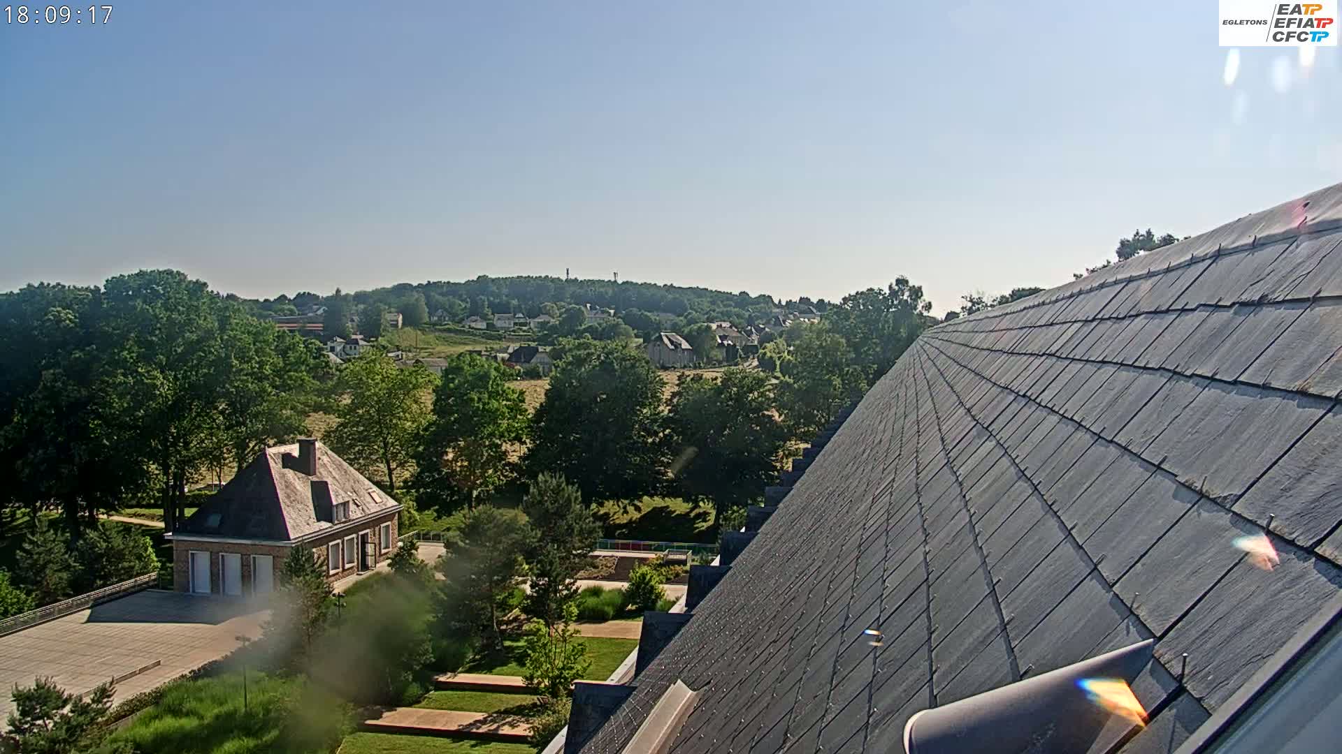 A steeply pitched slate roof overlooks a tree-lined landscape with buildings and a clear blue sky.