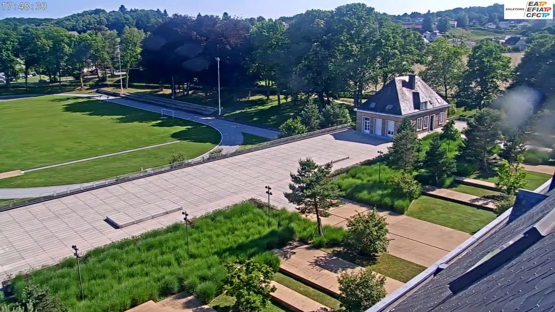An aerial view shows a grassy athletic field, paved areas, landscaping, and a small stone building on a sunny day.