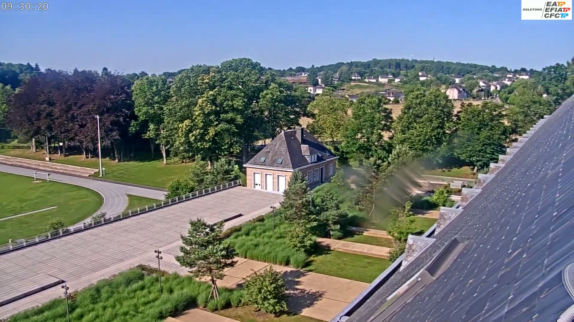 An elevated view shows a landscaped area with a small brick building, a running track, and a village in the background on a sunny day.