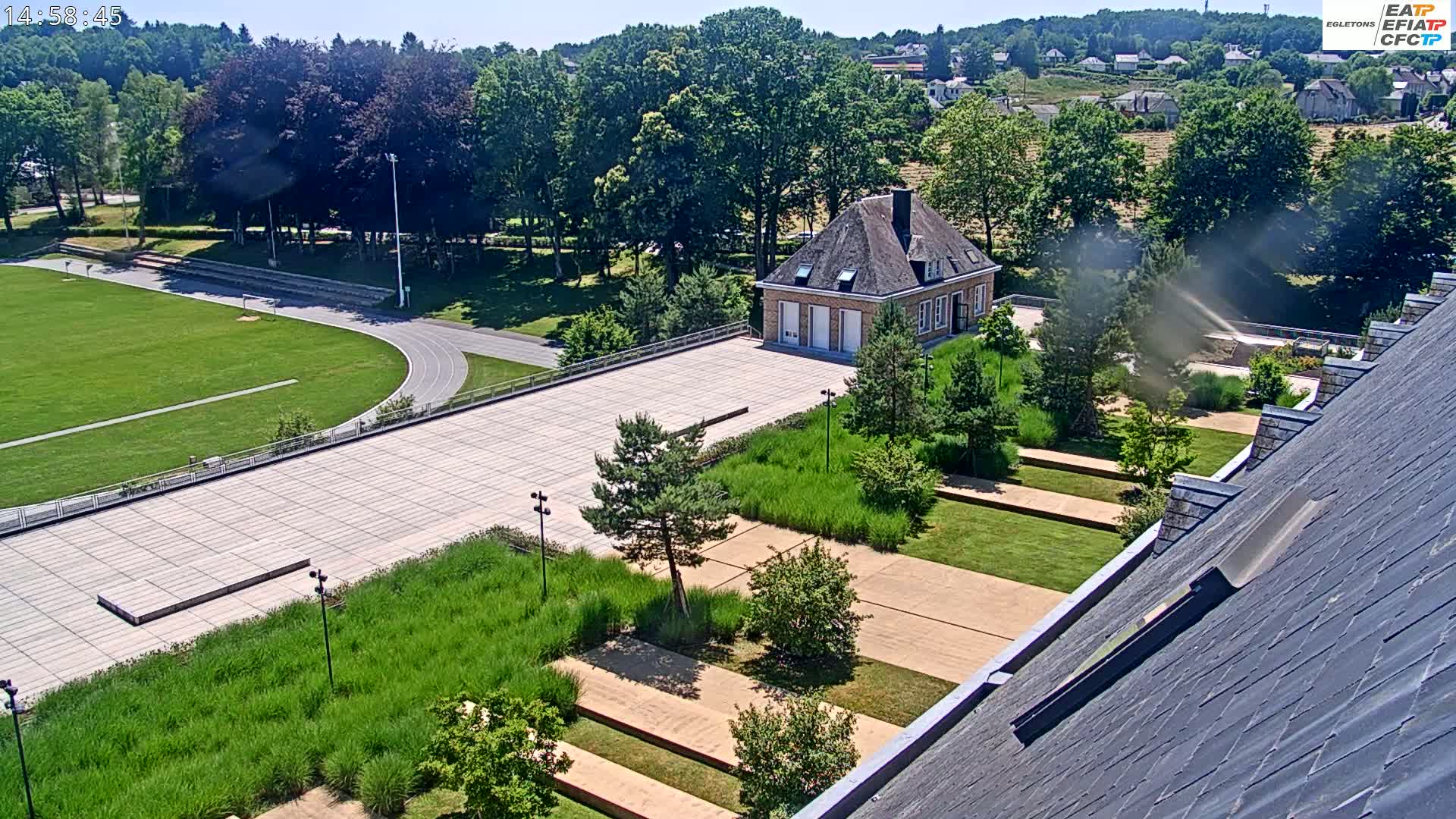 A high-angle view of a landscaped area with a running track, a small building, and a large paved plaza on a sunny day.