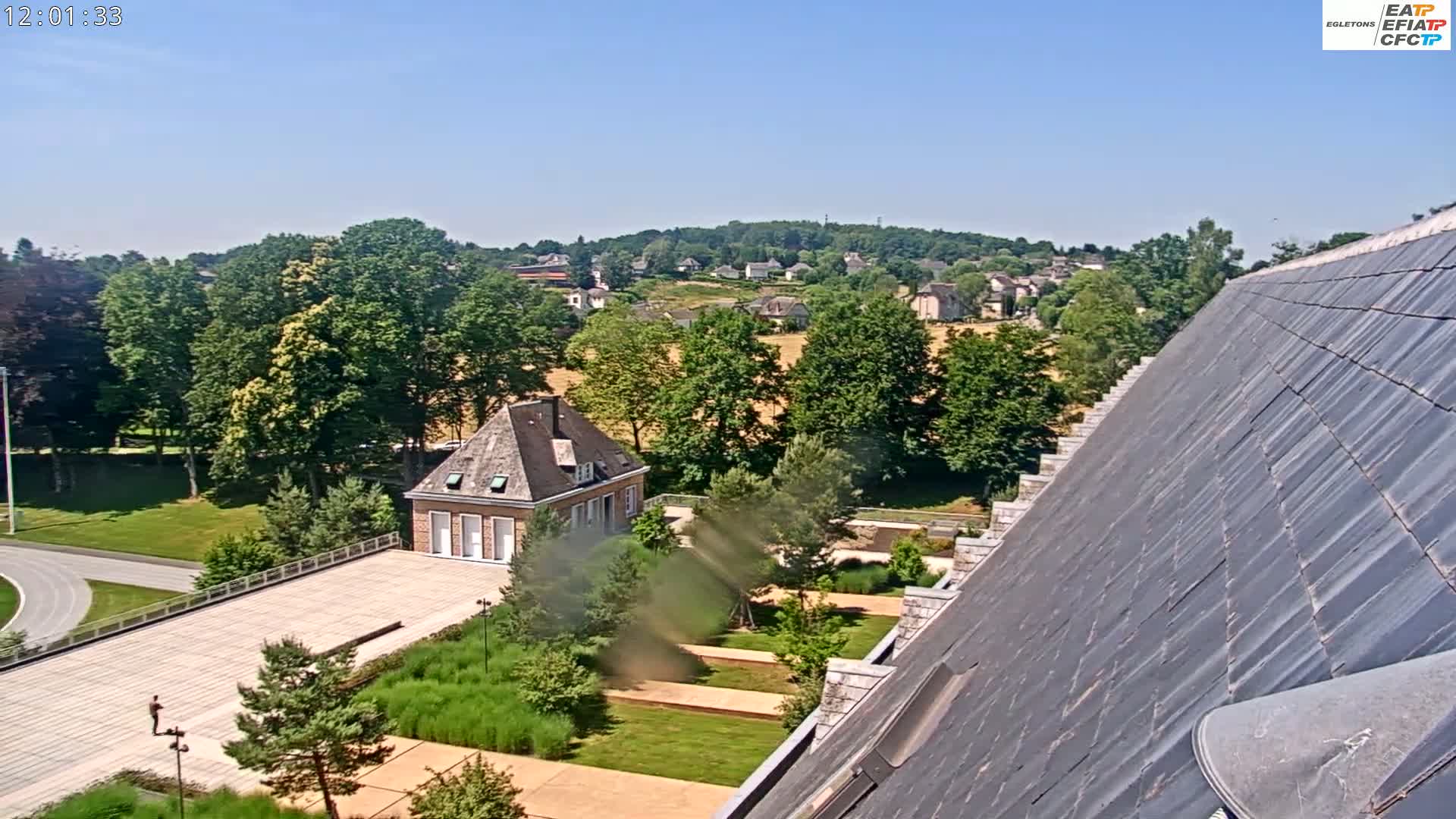 A sunny day reveals a high-angle view of a landscaped courtyard with a building, trees, and a small village visible in the distance.