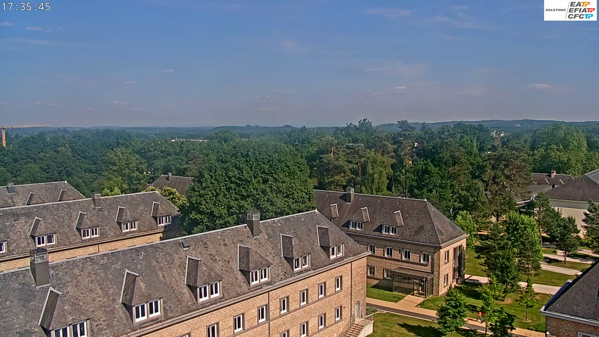 A high-angle view of several low-rise buildings with gray slate roofs, situated in a verdant landscape under a mostly clear sky.